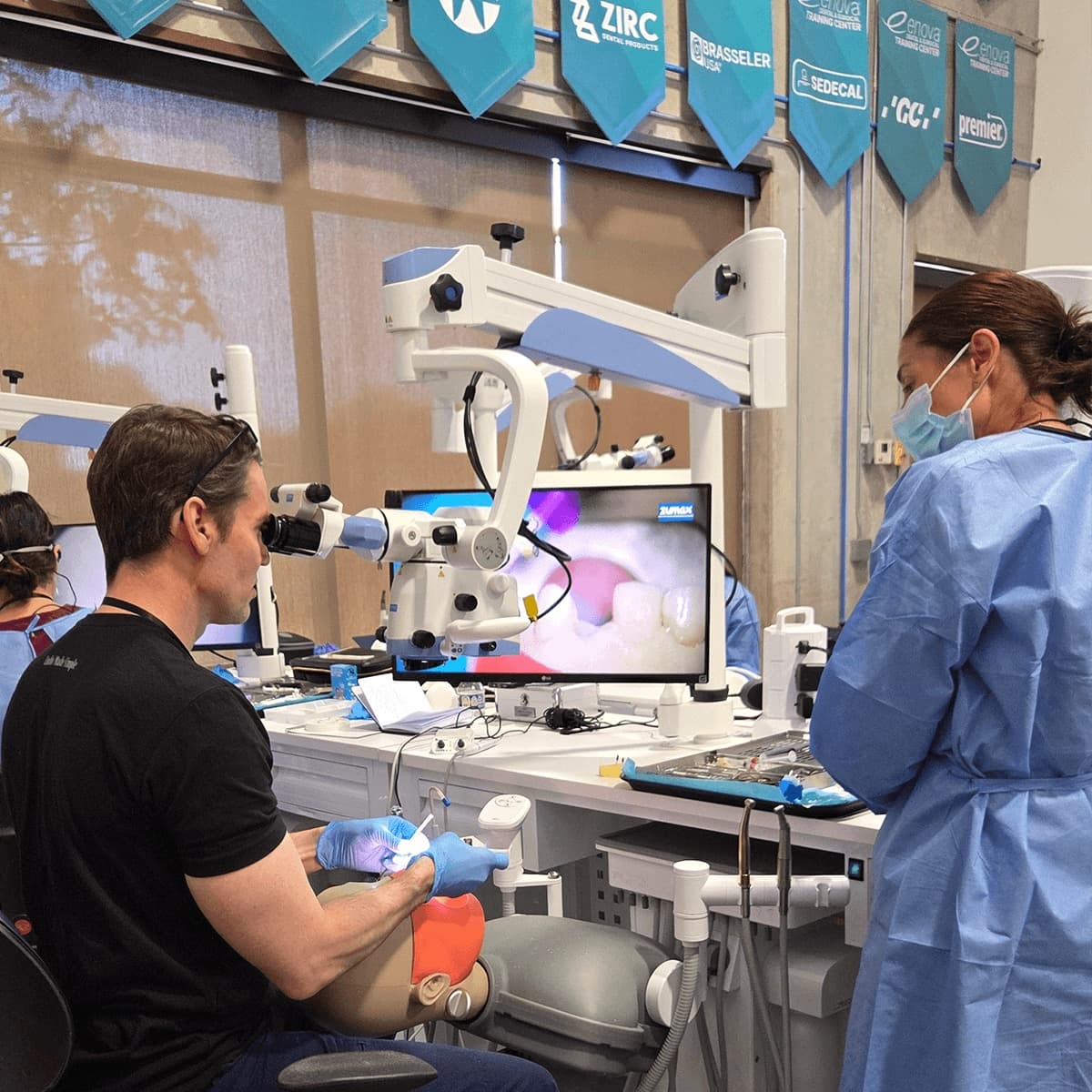 A workshop participant treating a manikin under the operating microscope with rubber dam in place, the magnified canal view live on the monitor beside him, while a gowned instructor watches over his shoulder