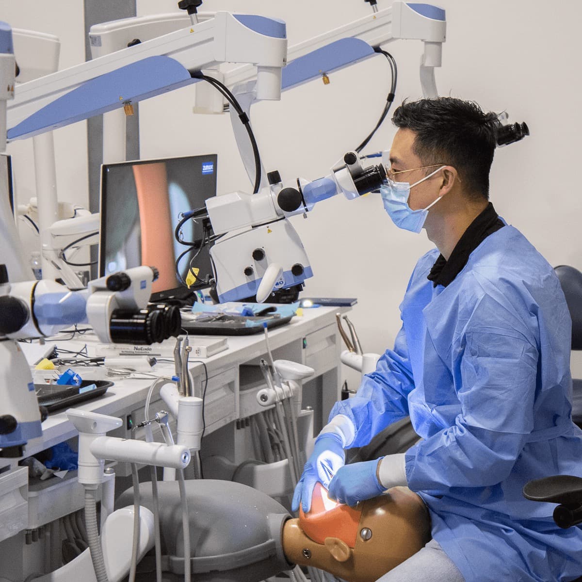 A participant leaning into the eyepieces with both gloved hands at the manikin, an instrument tray and the NuEndo textbook laid out on the bench
