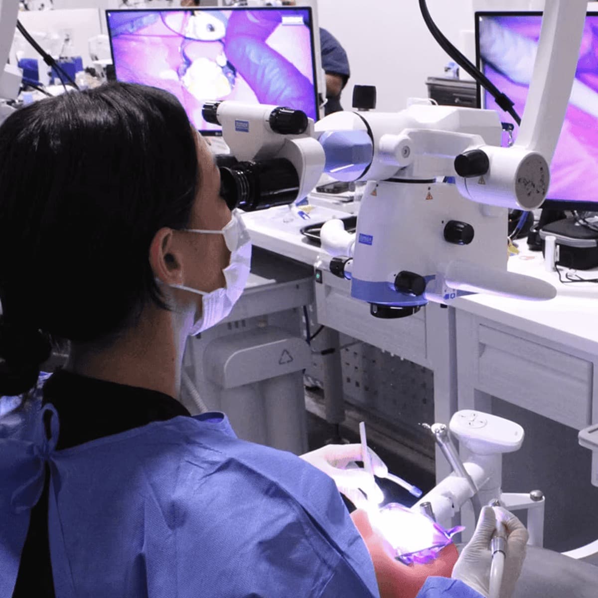 Over the shoulder of a participant at the scope, handpiece in gloved hand at the manikin, the magnified working view filling the monitor behind