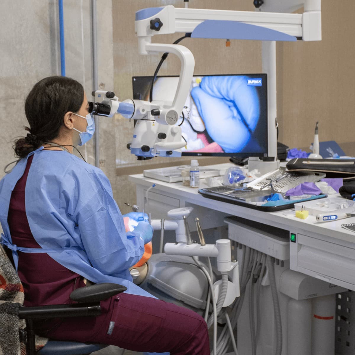 A participant in maroon scrubs at the eyepieces with a full instrument tray spread beside her, the monitor showing her magnified working view